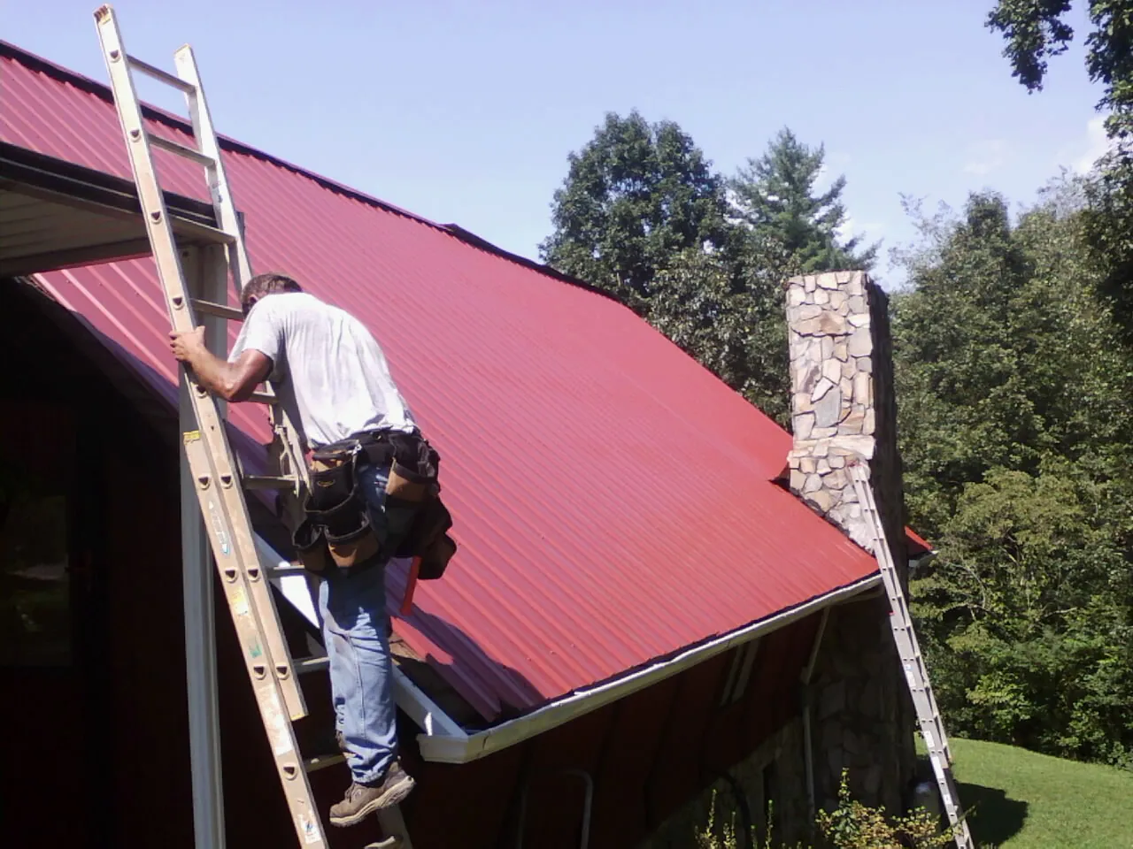 Roofer on a ladder beside a red metal roof and stone chimney — Soffit & Fascia Repair by Roofing Brothers in Asher